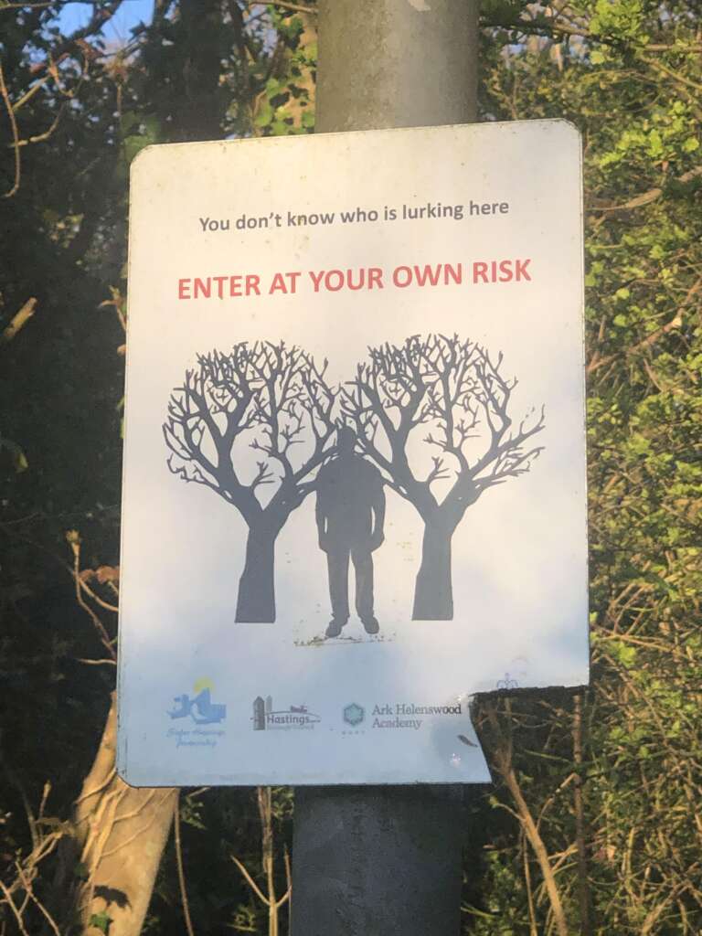 A warning sign on a pole shows an illustration of a person lurking between two trees shaped like eyes. The text reads, "You don't know who is lurking here. ENTER AT YOUR OWN RISK." Background shows dense trees and foliage.