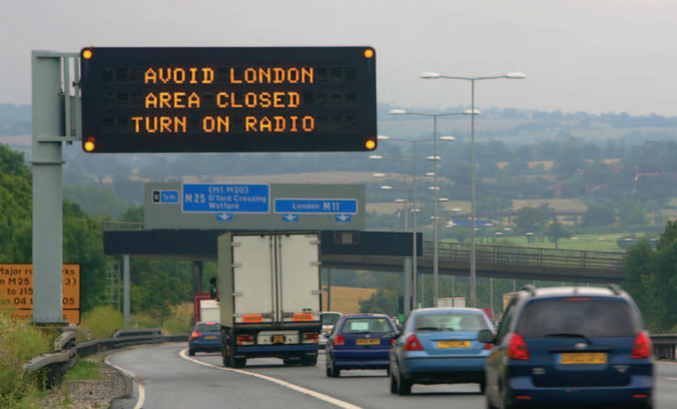 A digital road sign above a busy motorway advises: "AVOID LONDON. AREA CLOSED. TURN ON RADIO." Below, various vehicles drive on the road, with a distant landscape featuring hills and fields. Directional signs ahead indicate paths to Watford and London M11.