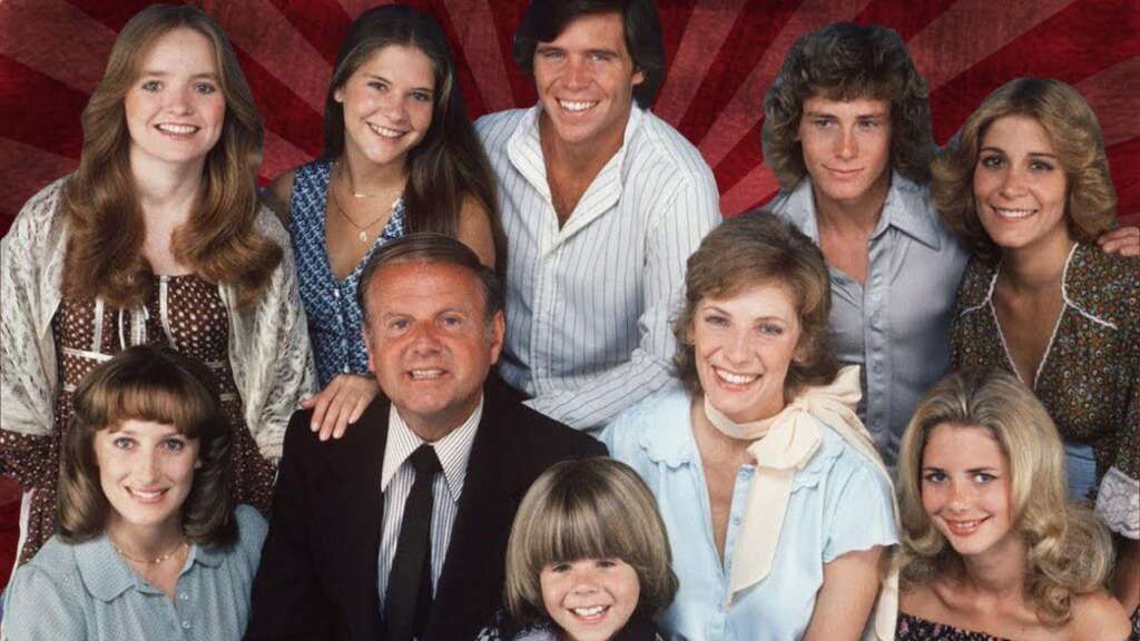 A group photo featuring eleven smiling people, including six women, four men, and one child. They are dressed in various styles of casual and semi-formal clothing. Many have 1970s-inspired hairstyles, and the background has a red radial design.