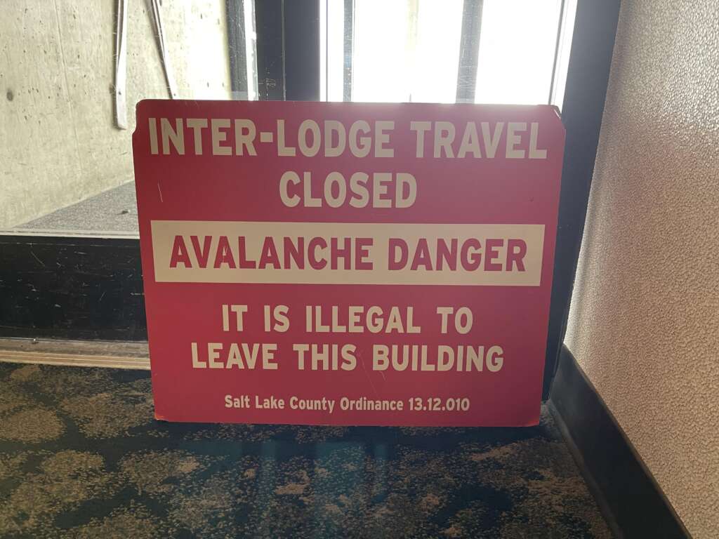 A red sign with white text reads: "INTER-LODGE TRAVEL CLOSED. AVALANCHE DANGER. IT IS ILLEGAL TO LEAVE THIS BUILDING. Salt Lake County Ordinance 13.12.010." The sign is placed inside a building near a glass door.