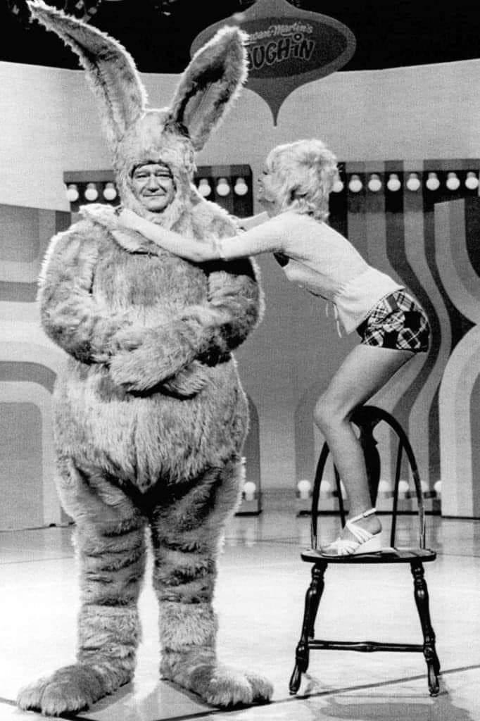 A man in a large, fluffy bunny costume stands on stage with a woman smiling and reaching up to embrace him. The woman is standing on a chair to reach the man's shoulders. The background features colorful, retro-style decor.