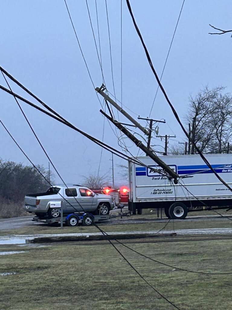 A damaged utility pole is leaning on power lines above a grey pickup truck towing a trailer, which is stopped near a white truck with red flashing lights. It's a cloudy day with wet ground and sparse trees in the background.