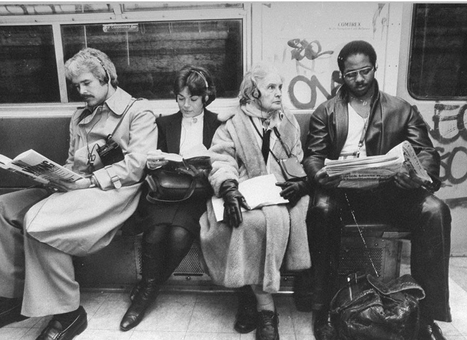 A black-and-white photo of four people sitting on a subway bench. The group consists of two men and two women, all engaged in reading newspapers or magazines. The subway car is marked with graffiti on the walls above their heads.