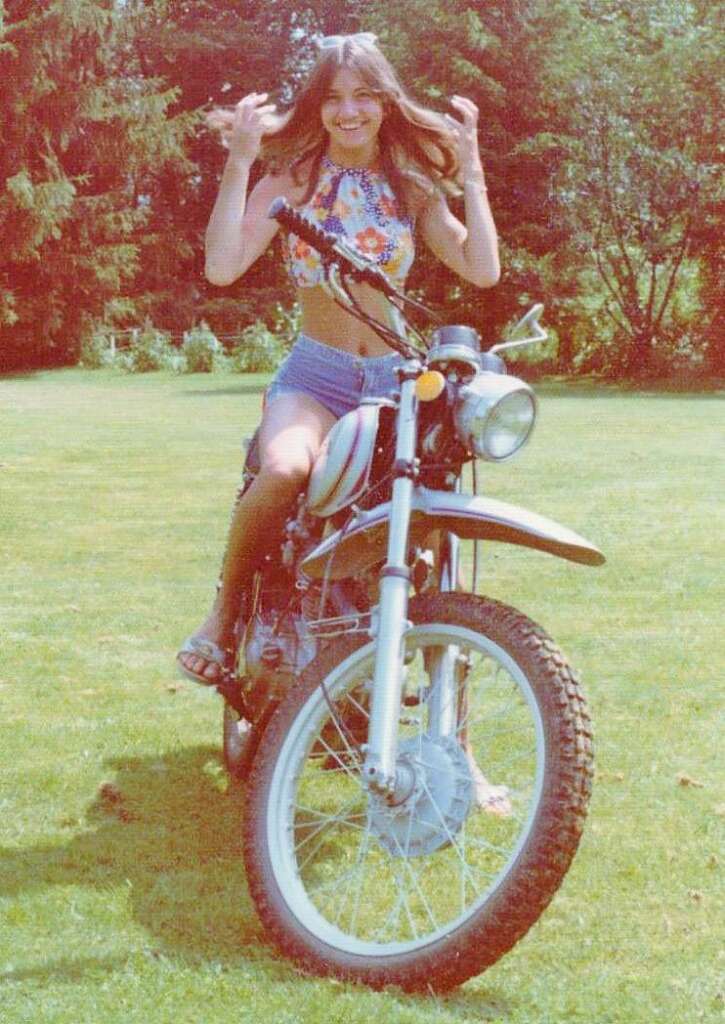 A person with long hair, wearing a floral crop top and denim shorts, is sitting on a vintage motorcycle in a grassy area with trees in the background. They are smiling and appear to be adjusting their hair.