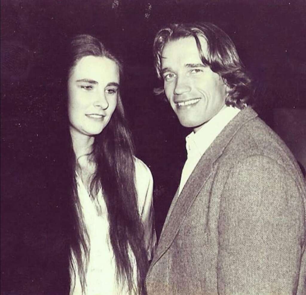 A man and a woman are standing side by side in this black and white photo. The man, dressed in a suit jacket and shirt, is smiling at the camera. The woman, with long hair and a white outfit, is looking slightly away from the camera.