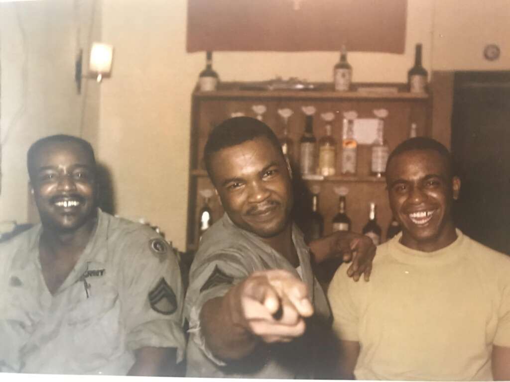 Three men are smiling and posing together in a room with a wooden bar and various bottles behind them. The man in the center is pointing towards the camera, while the other two are dressed in casual and military attire, respectively. The mood is jovial and relaxed.