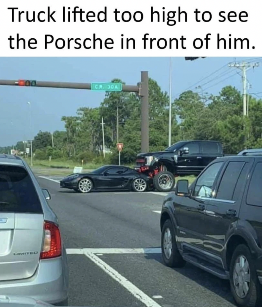 A lifted truck at an intersection has accidentally partially driven over the back of a small black Porsche sports car in front of it. There are other vehicles stopped at the intersection, and the scene is surrounded by greenery and some power lines. The caption reads, "Truck lifted too high to see the Porsche in front of him.