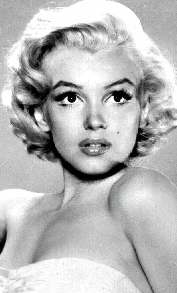 Black and white portrait of a woman with platinum blonde, wavy hair styled in a classic mid-20th-century fashion. Her expressive eyes and arched eyebrows stand out, and she has a beauty mark on her left cheek. She is gazing to the side, with light makeup and bare shoulders.