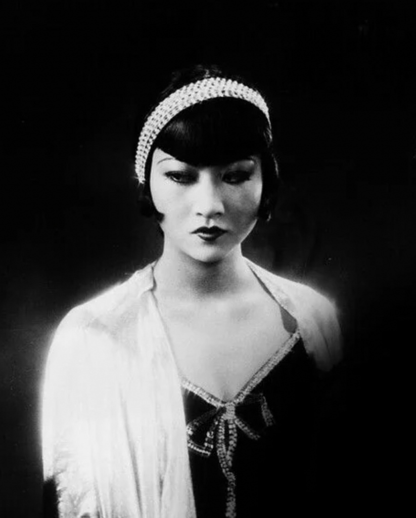 A black and white photograph of a woman dressed in 1920s fashion. She has a bob haircut with bangs and is wearing a headband with pearls. She has a serious expression and is dressed in elegant attire with a light shawl over her shoulders.