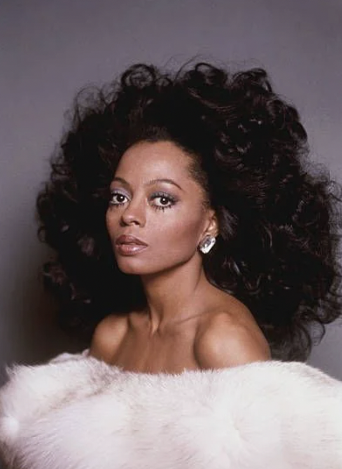 A woman with voluminous dark curly hair poses against a gray backdrop. She wears elegant earrings and a white fur wrap draped over her shoulders. Her expression is calm and poised, highlighting her radiant complexion and refined makeup.