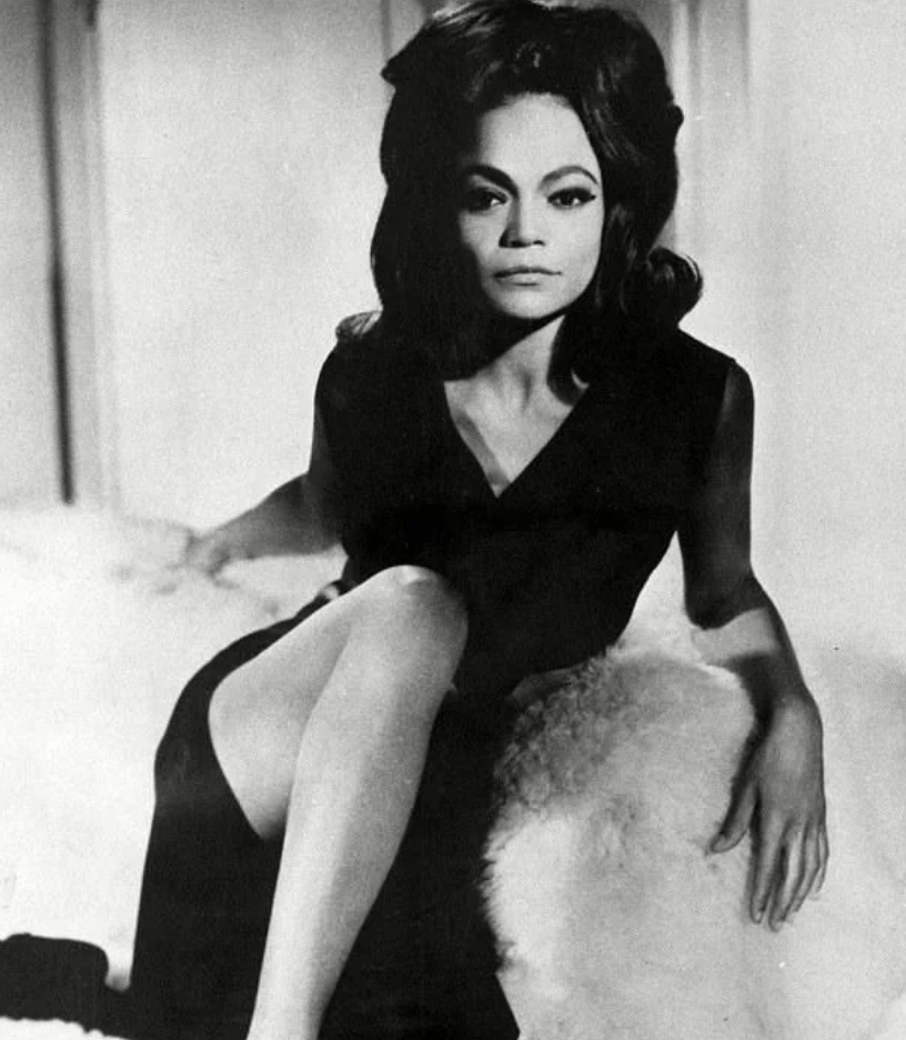 A black and white photo of a woman with voluminous dark hair, seated on a plush, fur-covered surface. She is wearing a sleeveless, dark-colored dress with a high slit, revealing one leg. The woman has a poised, confident expression and a striking pose.