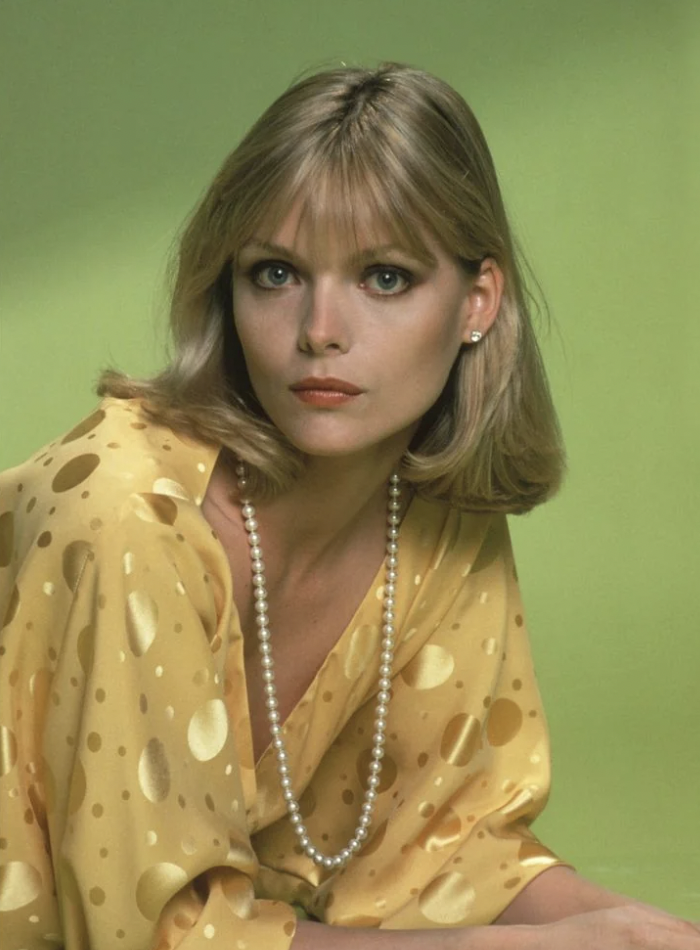 A woman with shoulder-length blonde hair and bangs is wearing a yellow blouse with polka dots and a pearl necklace. She has a serious expression and is set against a green background.