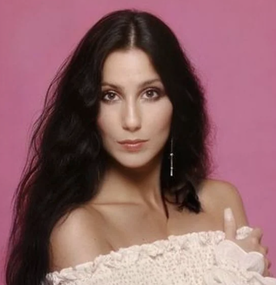 A woman with long, wavy dark hair poses against a pink background. She is wearing an off-the-shoulder white top and a single dangling earring. Her expression is neutral, and she gazes directly at the camera.