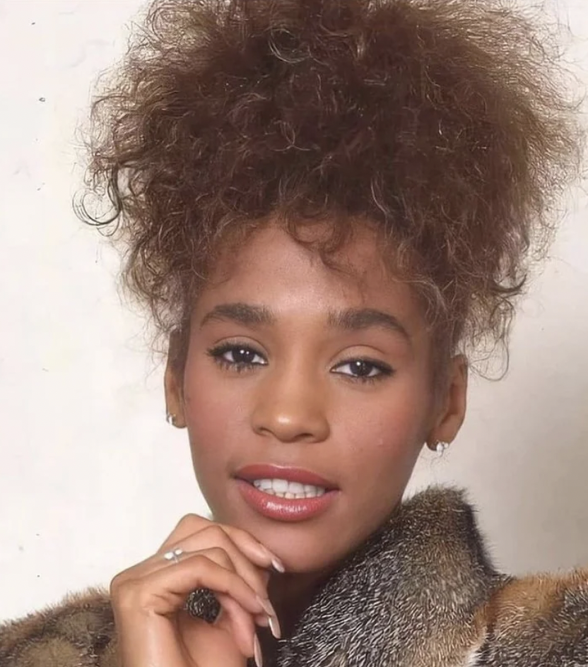 A woman with curly hair styled in an updo is pictured. She is wearing a fur coat, has subtle makeup with red lipstick, and is looking directly at the camera with a slight smile. She rests her chin gently on her hand, displaying a ring on her finger.