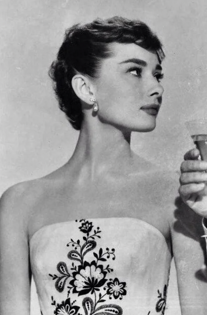 Black and white photo of a woman with short, dark hair styled elegantly, wearing a strapless dress adorned with an intricate floral pattern. She holds a small glass in her right hand and looks off to the side with a contemplative expression.