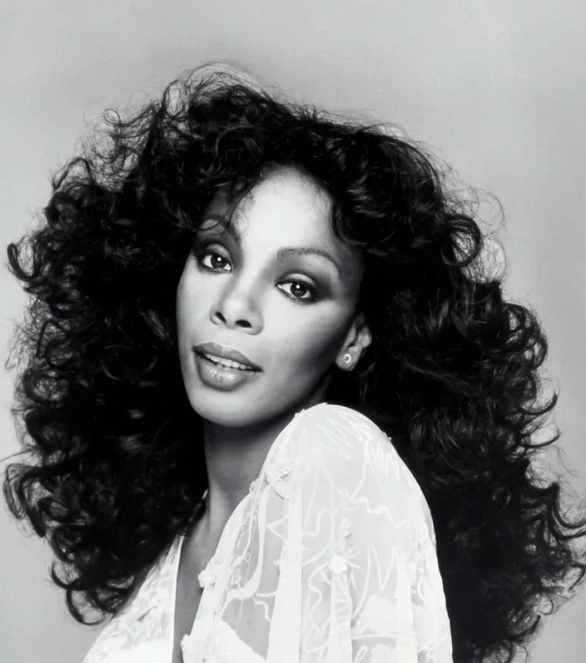 Black and white portrait of a woman with voluminous curly hair, wearing a sheer white lace top, looking directly at the camera with a calm expression. The background is plain, emphasizing her striking appearance.