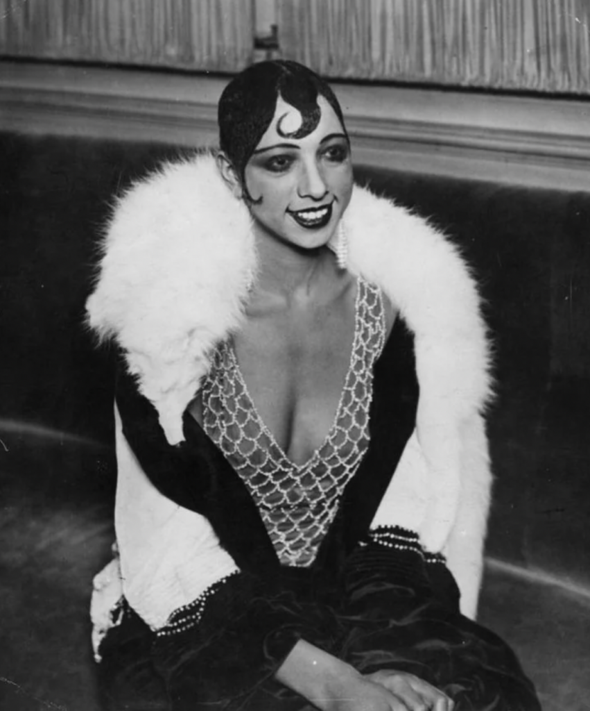 A woman dressed in 1920s fashion sits smiling. She wears a glamorous dress with a deep neckline, adorned with sparkling beads, and a white fur stole draped over her shoulders. Her short, sleek hair is styled with a prominent curl on her forehead.