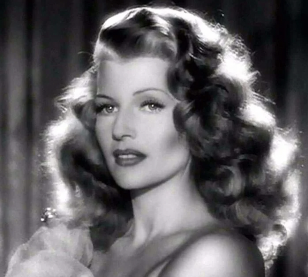 A black and white portrait of a woman with wavy, voluminous hair. She has a poised expression and is looking slightly to her right. The background is blurred, emphasizing her well-defined features and glamorous hairstyle.