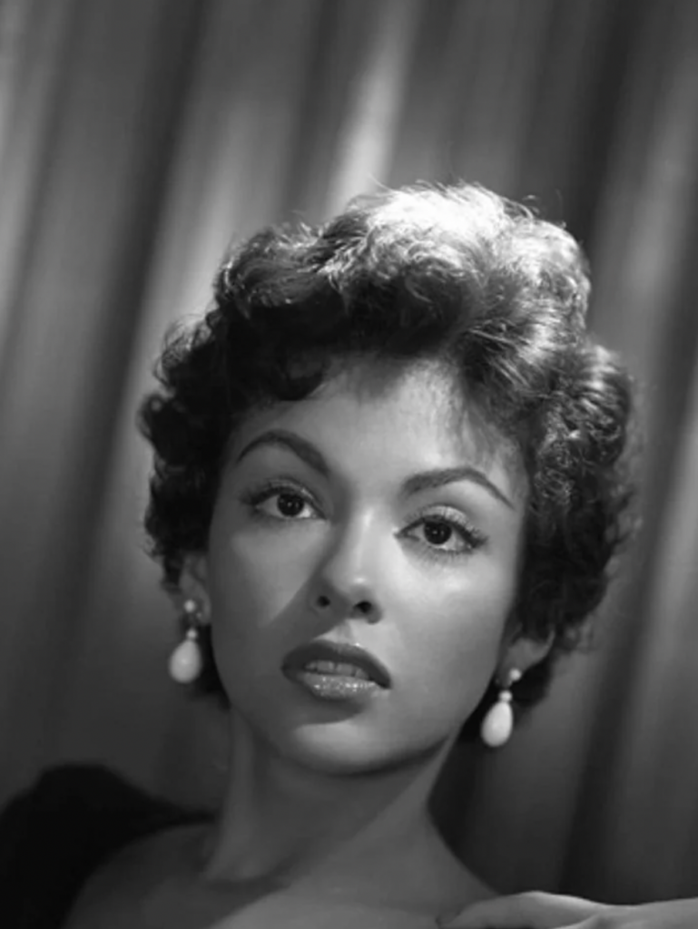 A black-and-white portrait of a woman with short curly hair, wearing drop earrings and a dark top. The background features draped curtains, and the lighting casts soft shadows on her face while highlighting her expressive eyes and elegant features.