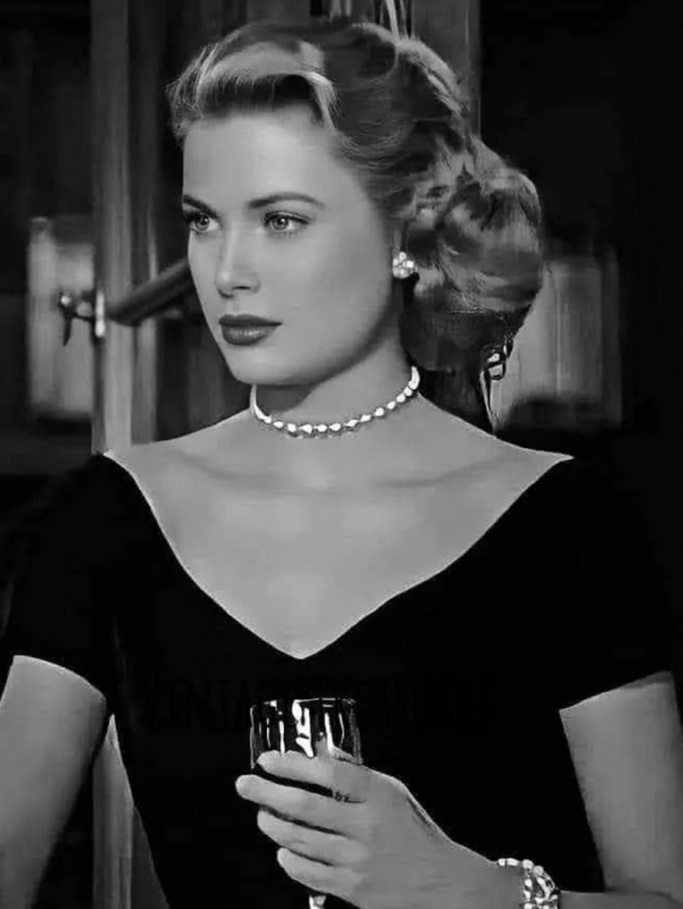 A woman with wavy blonde hair is dressed in an elegant off-the-shoulder black dress, accessorized with pearl earrings and a matching necklace. She is holding a wine glass and gazes thoughtfully into the distance, exuding a vintage, classic Hollywood glamour.