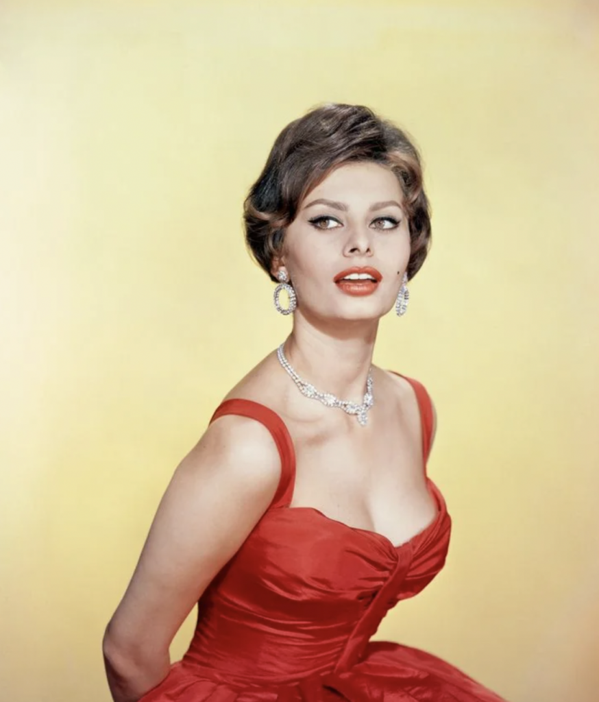 A woman with short brown hair, wearing a red sleeveless dress, a silver necklace, and matching earrings poses in front of a yellow background. She is looking slightly up and to the side, with a confident expression.