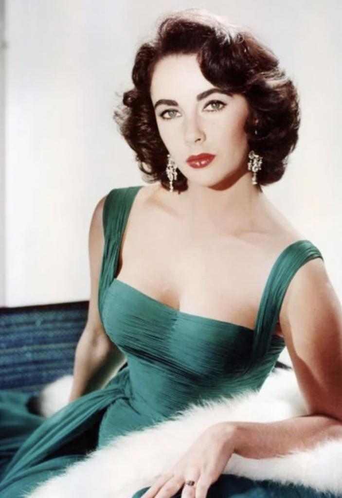 A woman with dark, wavy hair and red lipstick poses elegantly in a green, off-the-shoulder dress. She is wearing dangling earrings and has a confident expression. The background is softly lit, highlighting her glamorous appearance.