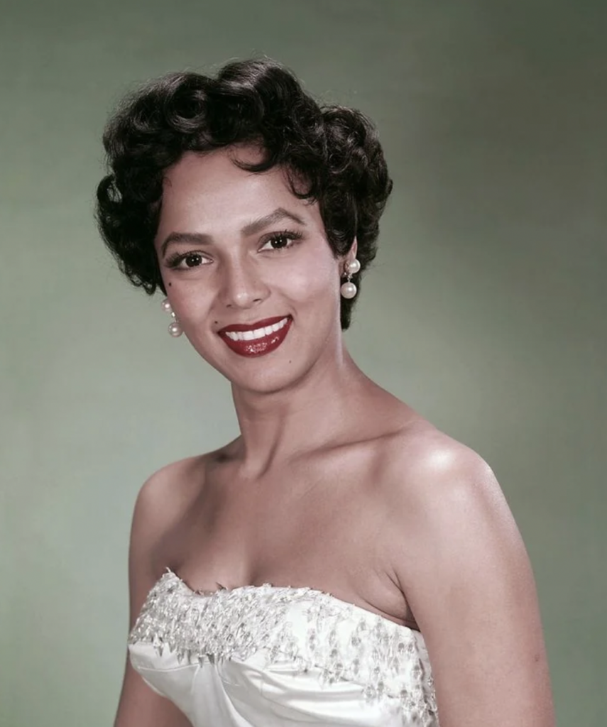 A woman with short, curly dark hair smiles at the camera. She is wearing pearl earrings and a strapless white dress with intricate, beaded detailing on the bodice. The background is a gradient of soft green hues.