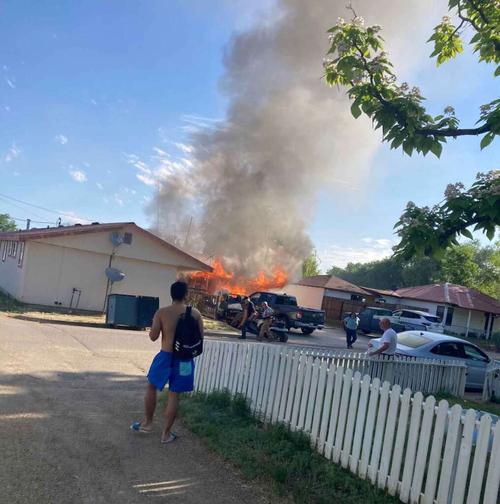 A fire engulfs a residential building, sending thick smoke into the sky. A shirtless person in blue shorts stands in the foreground, holding a bag. Several other individuals observe and react to the fire. A white picket fence and trees are visible in the scene.