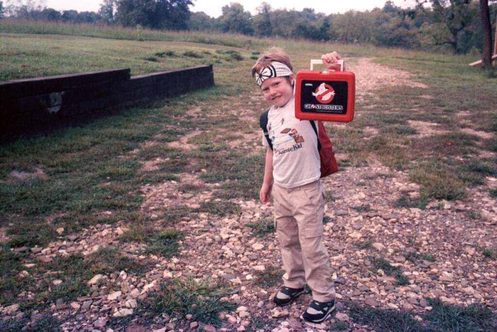 A young boy stands outdoors on a rocky path holding a red Ghostbusters lunchbox. He is wearing a white headband, a light-colored shirt with a graphic, khaki pants, and sneakers. The background features a grassy area with trees and a retaining wall.