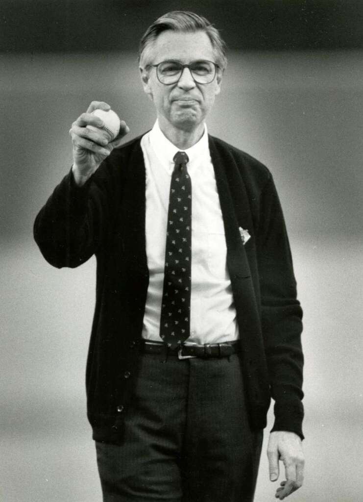 A man wearing glasses, a patterned tie, and a dark cardigan holds a baseball in his right hand. The background is blurred, suggesting an outdoor setting. The man appears to be standing still and looking directly at the camera.