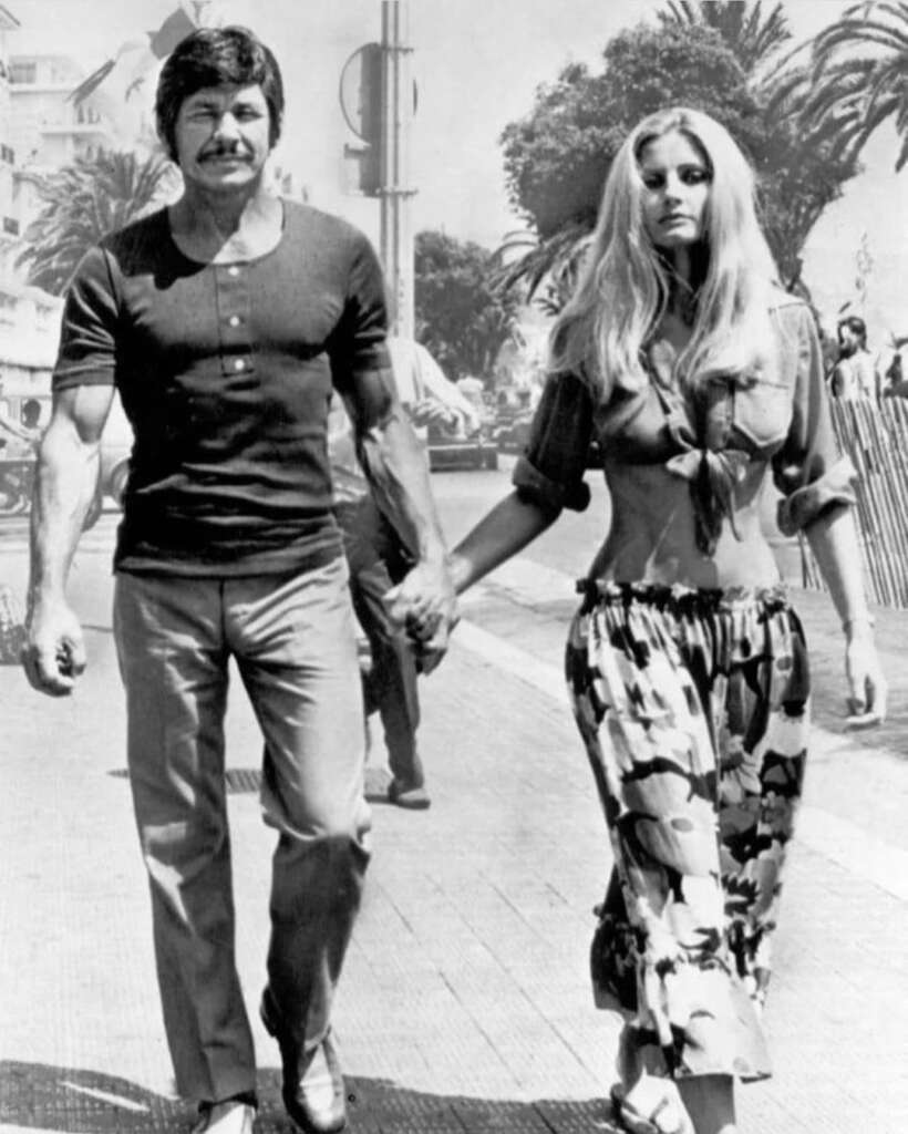 A couple holds hands while walking outdoors. The man has a mustache, wears a T-shirt, and slacks. The woman has long blonde hair, wears a crop top tied at the waist, and patterned pants. Palm trees and people are visible in the background.