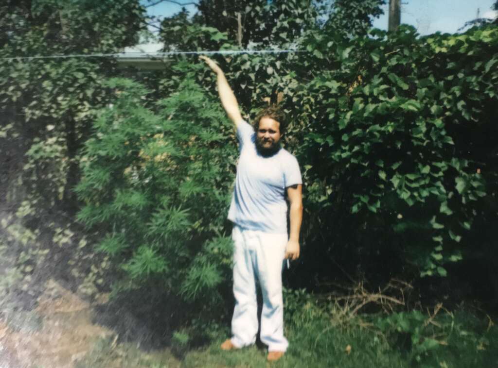 A person with a beard, wearing a light-colored shirt and pants, stands barefoot in a garden with lush green foliage. They are standing next to a tall plant, reaching up with one arm to touch a string or line above them.