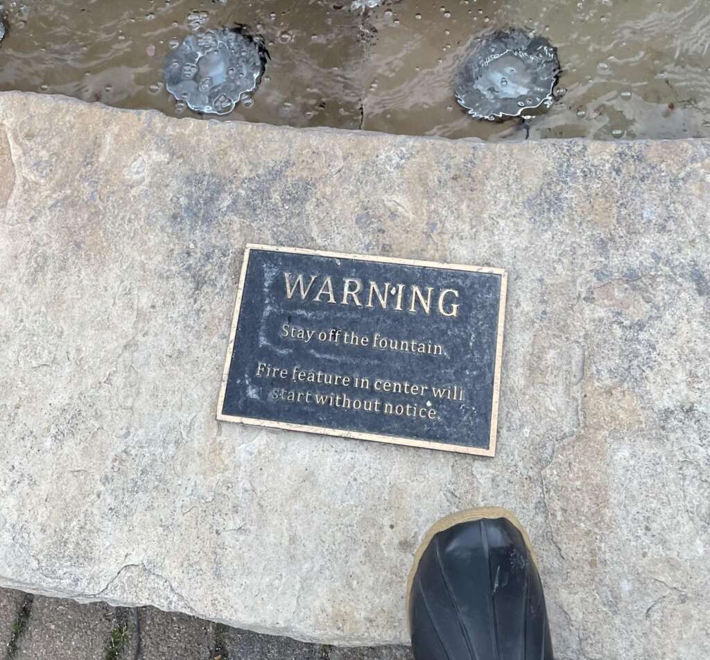 A warning sign is displayed on a stone ledge next to a fountain. The sign reads, "WARNING: Stay off the fountain. Fire feature in center will start without notice." A portion of a person’s black shoe is visible at the bottom right corner of the image.