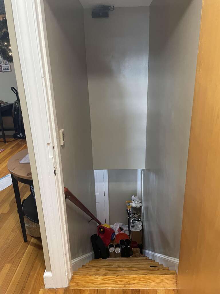 A view looking down a narrow staircase with clutter at the bottom, including boxes, a bucket, and various household items. The walls are light gray, and the wooden steps are visible. The staircase railing is on the left side.