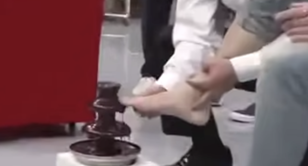 A person in a white shirt is holding and dipping a foot into a chocolate fountain. Another person dressed casually is sitting nearby. The scene appears to be taking place indoors, with a red object partially visible in the background.