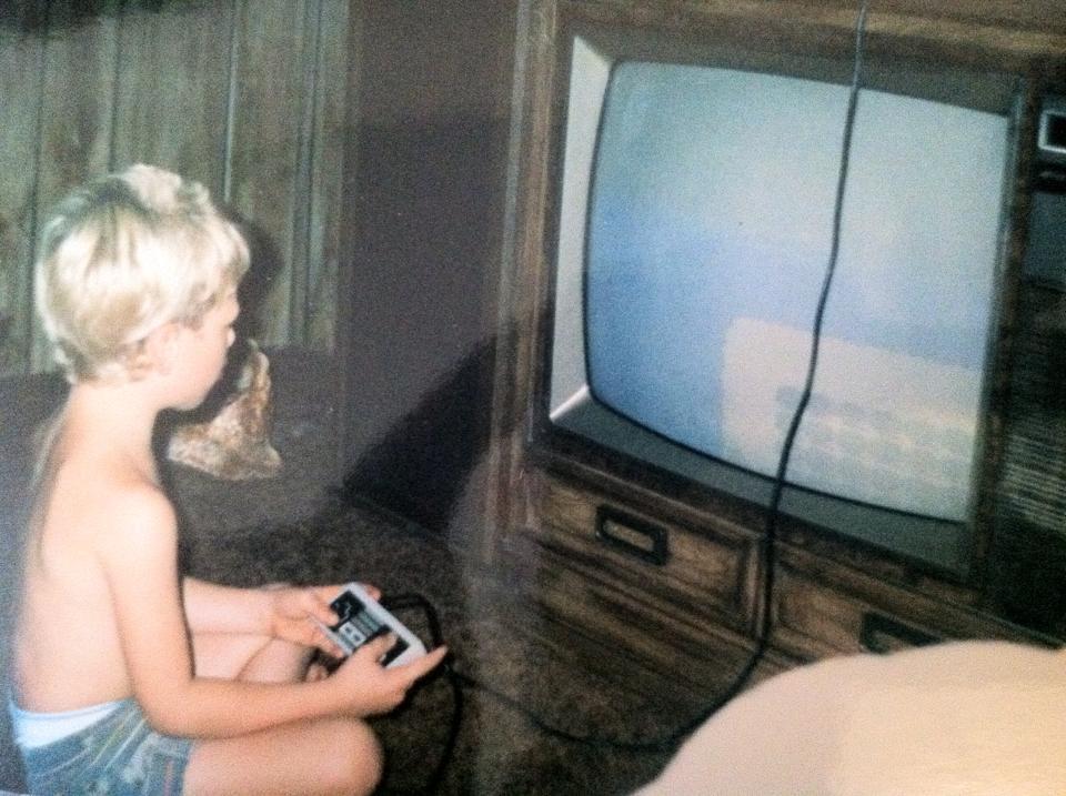 A young child with blond hair is sitting on the floor, holding a classic video game controller. They are facing an old-fashioned, boxy television with a blank screen. The scene appears to be from the 1980s or 1990s, in a room with wooden paneling.