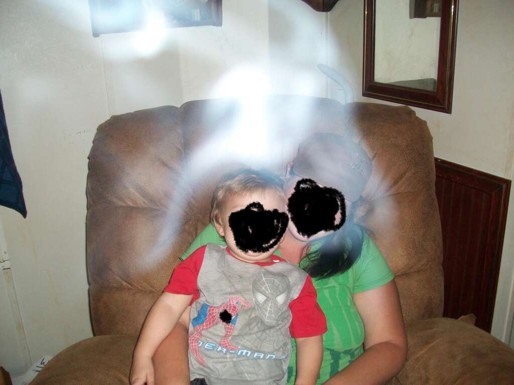 A young child and an adult, whose faces are obscured, sit together in a brown armchair. The child is wearing a red and gray Spiderman shirt, and the adult is dressed in a green shirt and cap. A bright, mist-like light appears above and around them, creating a ghostly effect.