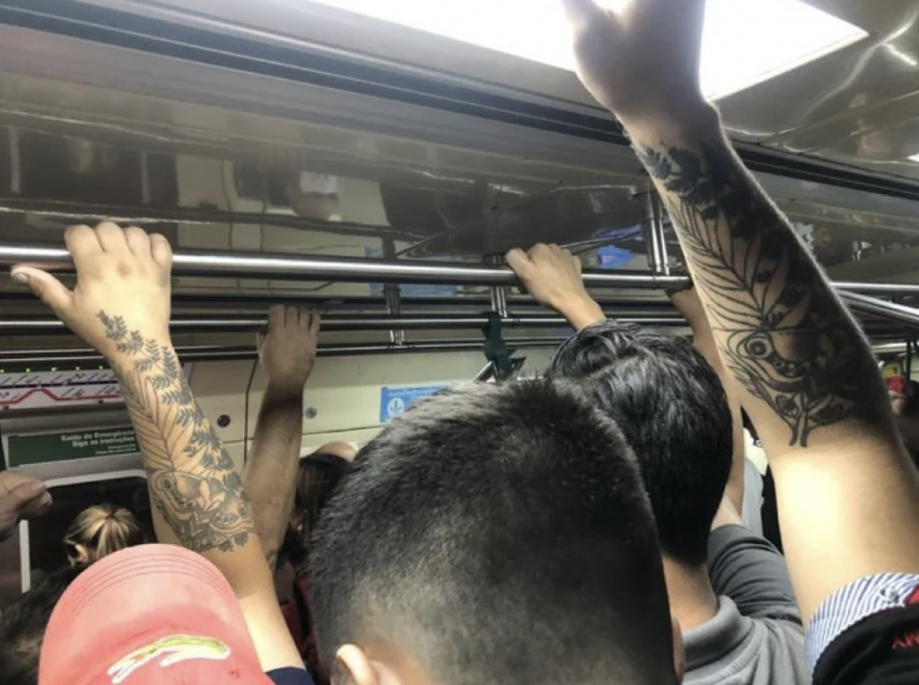 A group of people ride in a crowded subway car, with many holding onto overhead handrails. Several visible arms feature various tattoos, including designs of feathers, flowers, and an owl. A red cap and short dark hair are noticeable.