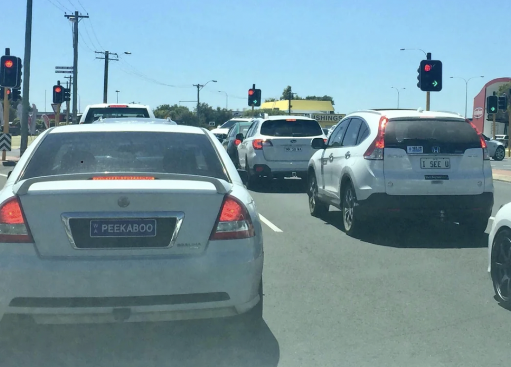 A line of cars is stopped at a red light on a sunny day. Two notable license plates are visible: the car on the left has a plate reading "PEEKABOO," and the car on the right has a plate reading "I SEE U." The sky is clear, and traffic lights are red.