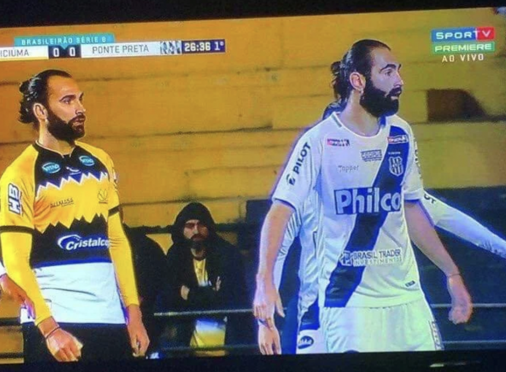 Two soccer players are on the field during a Brazilian league match between Criciúma and Ponte Preta. The player on the left wears a yellow and black uniform, and the player on the right wears a white and black uniform. A spectator is seen in the background.