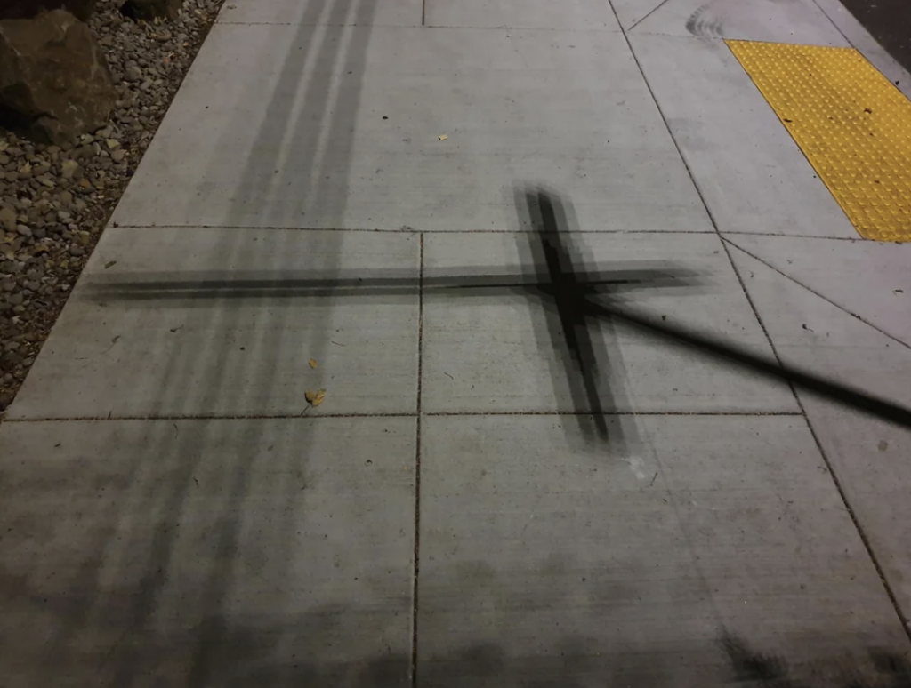 A nighttime view of a concrete sidewalk with black tire skid marks forming an "X" pattern. A patch of yellow truncated dome tiles is visible in the upper right corner, indicating a tactile paving surface for visually impaired pedestrians.