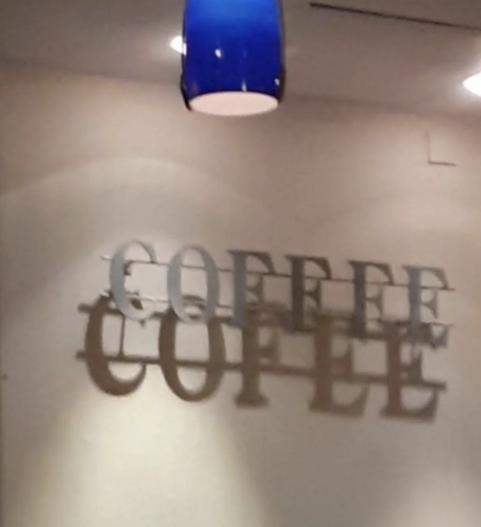 A wall with the word "COFFEE" is displayed twice in metal letters, creating a shadow effect. A blue light fixture hangs from the ceiling, casting light onto the wall, enhancing the shadow. The setting appears to be indoors, possibly at a café or coffee shop.