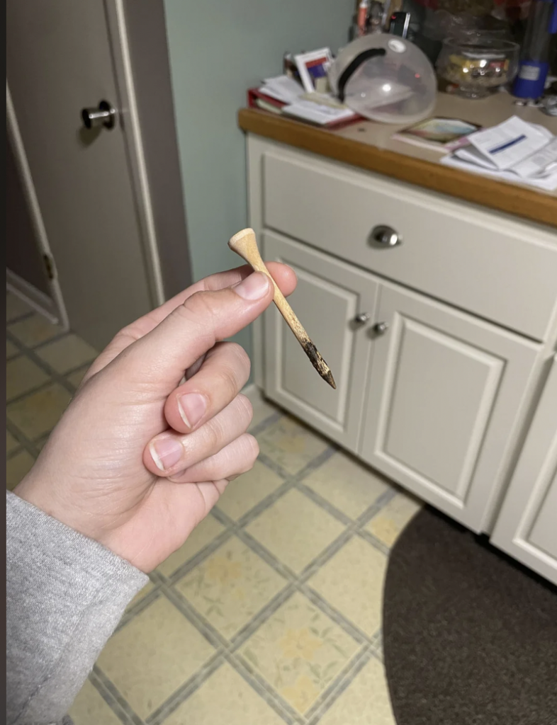 A hand is holding a small wooden item with a charred end, resembling a golf tee, over a kitchen floor with light-colored tiles. In the background, there's a white cabinet with a countertop cluttered with various items.