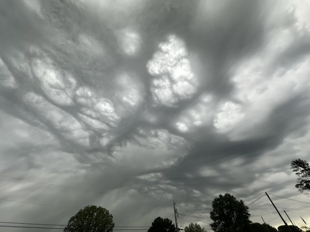 Dramatic sky with dark, swirling cloud formations and patches of blue sky peeking through. The view is framed by silhouettes of trees and utility poles below, creating a contrast between the turbulent sky and the calm landscape.