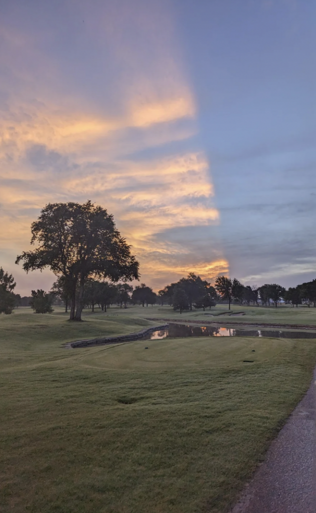 A serene landscape at dawn with a golf course featuring a small pond reflecting the morning light. The sky is painted with hues of orange and blue, and a striking shadow divides the clouds. Trees and grassy fields create a peaceful atmosphere.