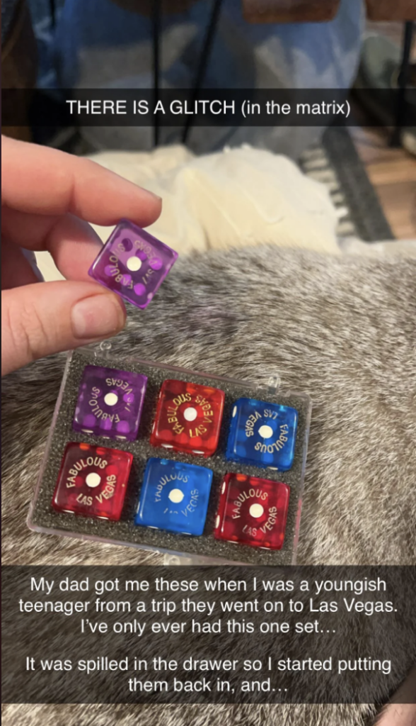 A hand holds a purple die with white dots. Below, there's a tray with six dice in purple, red, and blue, all labeled "Fabulous Las Vegas." The background shows a gray textured surface. Text overlays the image, describing the dice as a gift from a trip to Las Vegas.