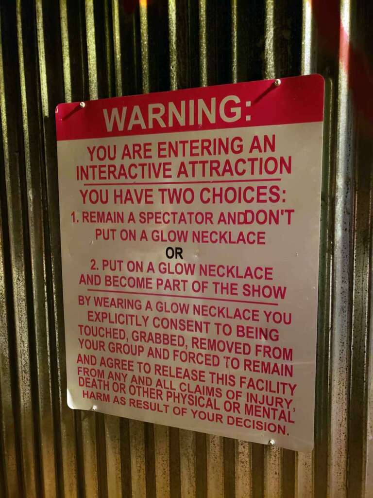 A sign with a bright red "WARNING" message against a metal wall. The sign reads: "You are entering an interactive attraction. You have two choices: 1. Remain a spectator and don't put on a glow necklace, or 2. Wear a glow necklace and become part of the show..." and continues explaining terms of participation and consent.