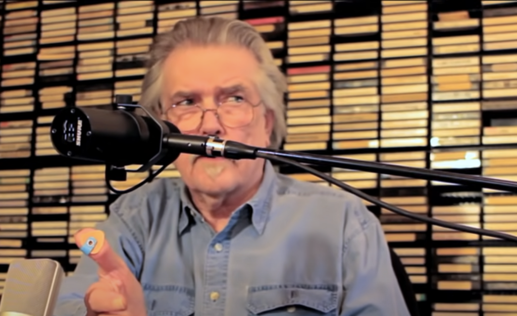 An older man with gray hair and a mustache is sitting in front of a microphone, holding a small, round object with a single eye illustration. He is wearing a denim shirt. The background consists of a wall filled with shelves of cassette tapes.