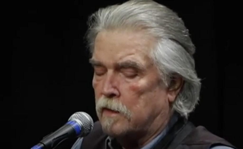 An older man with gray hair and a mustache is singing into a microphone. His eyes are closed, and he appears to be absorbed in his performance. The background is dark.