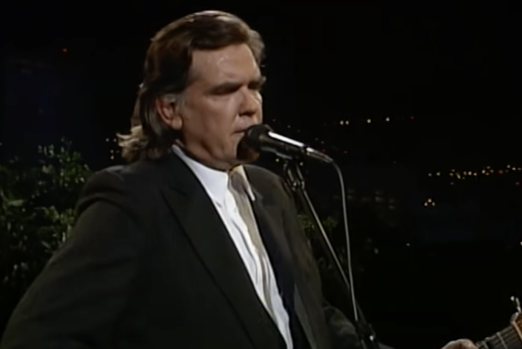 A person with long hair, dressed in a black suit and white shirt, stands in front of a microphone, playing a guitar. The background is dark with some lights visible in the distance. The person appears to be singing or speaking into the microphone.